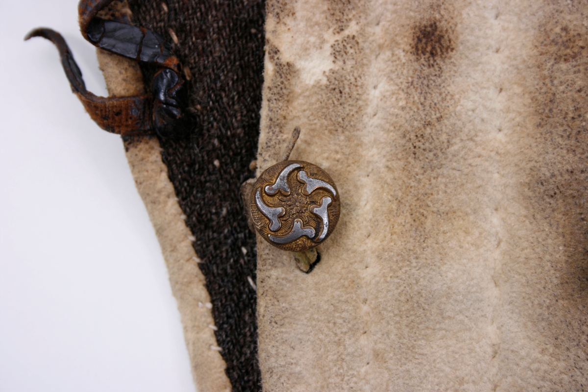 Håndsydd knebukse i lyst skinn. Lav midjeplassering. Linningen i midjen og lokket lukkes med ulike og graverte messingfargede knapper.  I høyre side ved linningen er det innsydd en lomme. I linningen MB er det splitt som lukkes med skinnsnor gjennom 3 x 2 hull og MF lukkes den med 3 knapper. Linningen er foret med ullstoff og har 4 rader prikkestikning gjennom for og skinn. Stor vidde over baken. Nede ved knærne er det splitter som lukkes med 4 knapper. Påsydd knelinning som trolig har hatt spenne for lukking. Alle knappene er festet med skinnsnor som igjen er festet på innsiden av plagget.Alle sammensyingskanter har tittekanter.