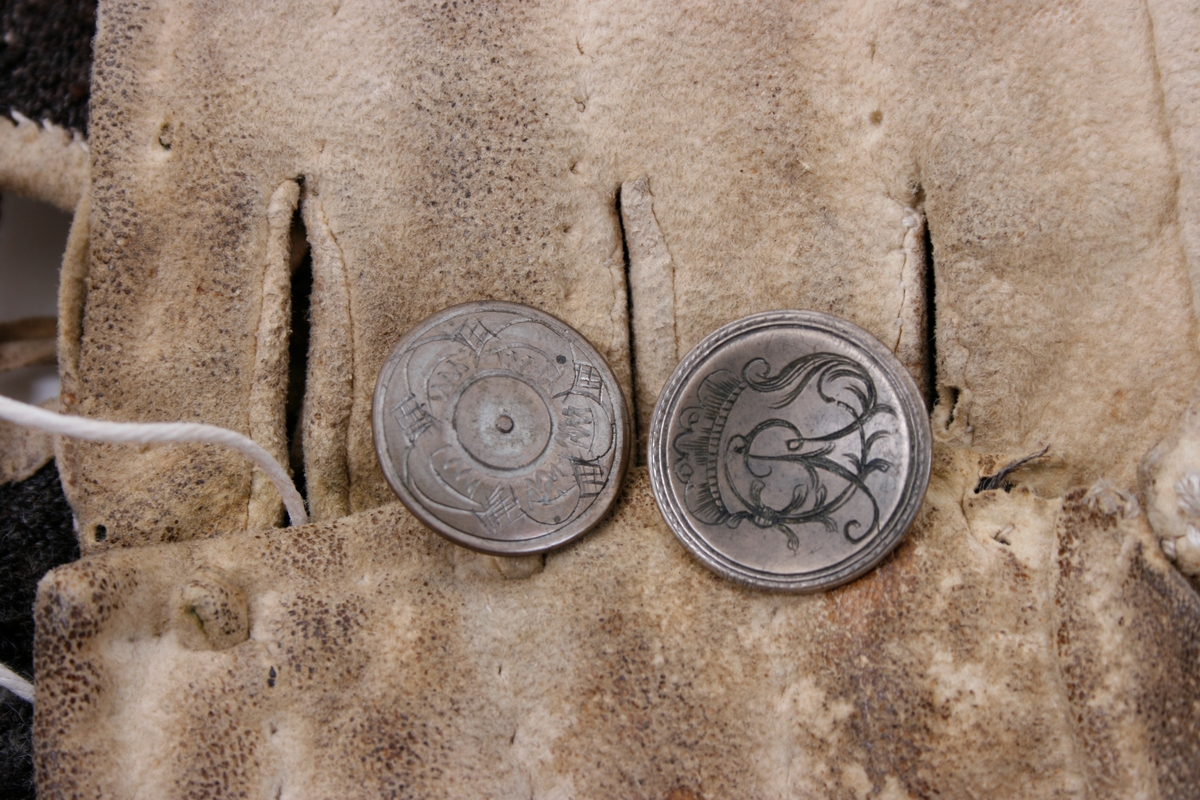 Håndsydd knebukse i lyst skinn. Lav midjeplassering. Linningen i midjen og lokket lukkes med ulike og graverte messingfargede knapper.  I høyre side ved linningen er det innsydd en lomme. I linningen MB er det splitt som lukkes med skinnsnor gjennom 3 x 2 hull og MF lukkes den med 3 knapper. Linningen er foret med ullstoff og har 4 rader prikkestikning gjennom for og skinn. Stor vidde over baken. Nede ved knærne er det splitter som lukkes med 4 knapper. Påsydd knelinning som trolig har hatt spenne for lukking. Alle knappene er festet med skinnsnor som igjen er festet på innsiden av plagget.Alle sammensyingskanter har tittekanter.