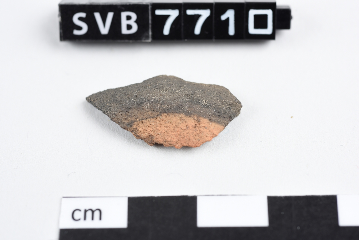 Savkt buet keramikkfragment. Glatt på innsiden, ru på utsiden. Formen er kantet og uregelmessig. Fargen er gråbrun med en rødbrun flekk.