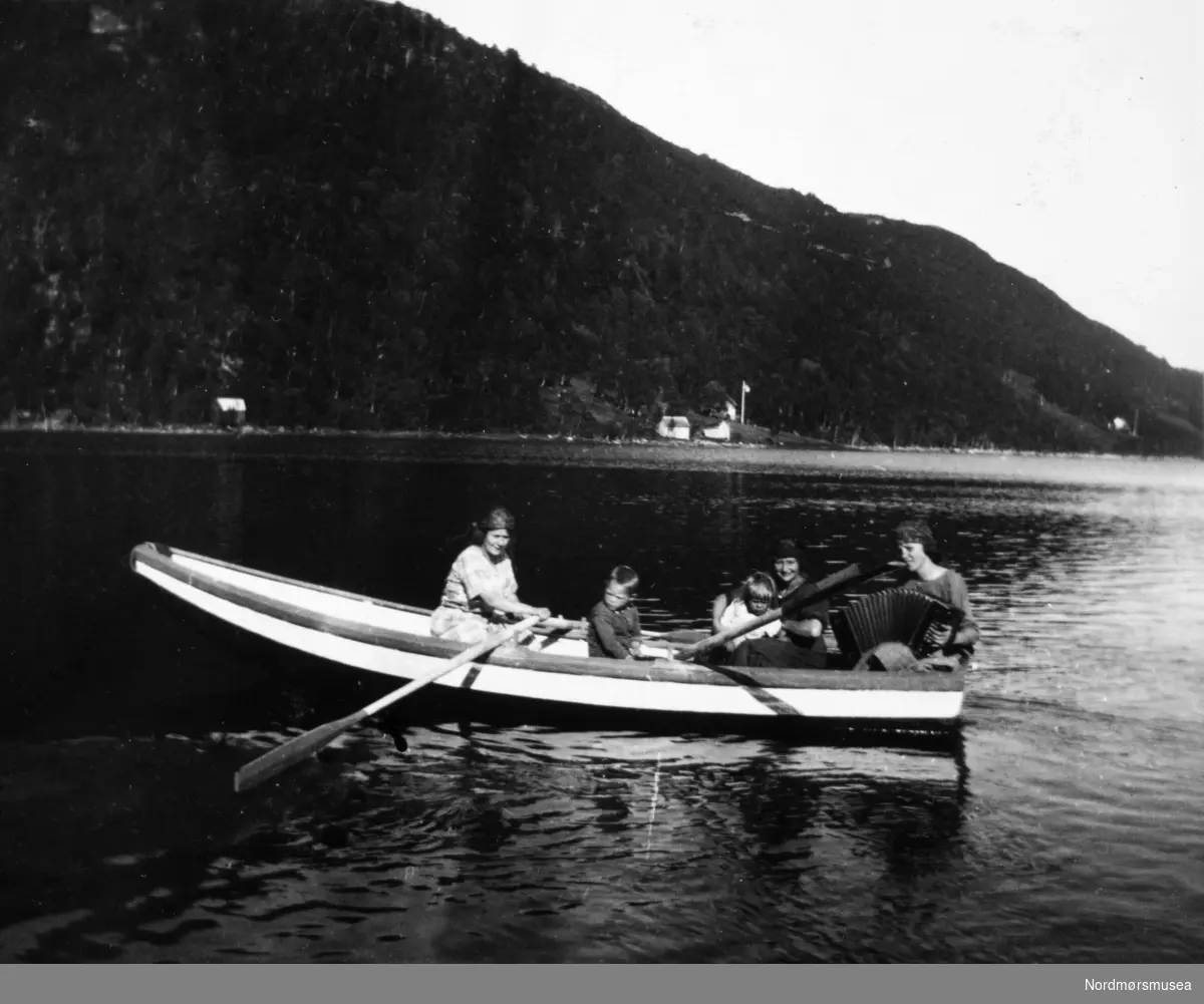 Tre kvinner og to barn ute på fjorden i robåt. Legg også merke til kvinnen bak med trekkspillet. Det er ukjent kvem vi ser på bildet, men de er trolig fra Williams-familien/slekten. Det er også ukjent hvor og når bildet er tatt. Fra et fotoalbum tilhørende Ellinor Williams-Phakdikun (1905-1963). Fra Nordmøre museums fotosamlinger.