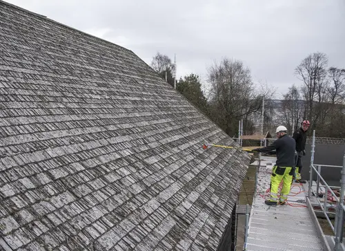 Låvetak med gamle takfliser blir børstet av menn med hjelm som står på stillas.