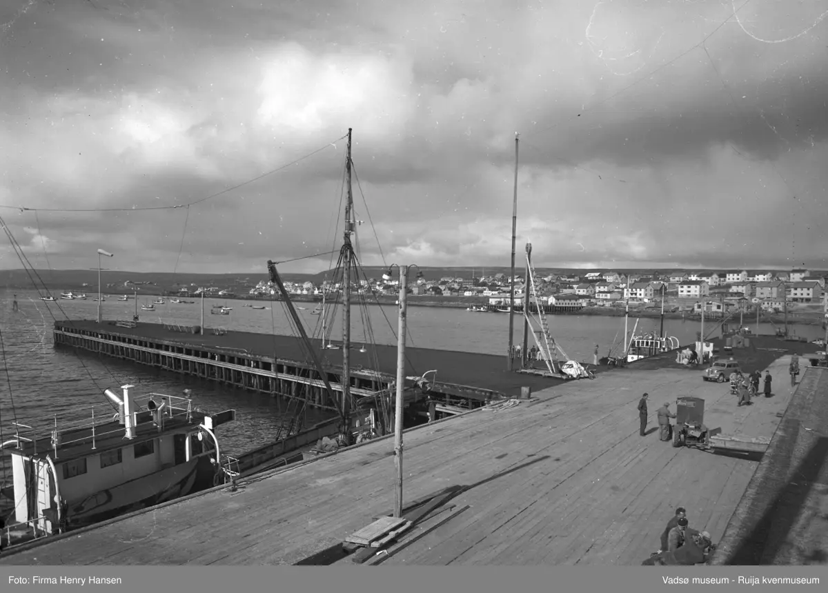 Vadsø 1952. Dampskipskaia. Bildet er tatt mot nordøst og viser Midtbyen og Ytrebyen, med overskyet himmel. Kaien er i forgrunnen, både landsiden og utstikkeren. To båter ligger ved kai på landsiden, nærmest ser vi mast og en del av baug og styrhuset. Det er folk på kaia, en truck og en personbil.