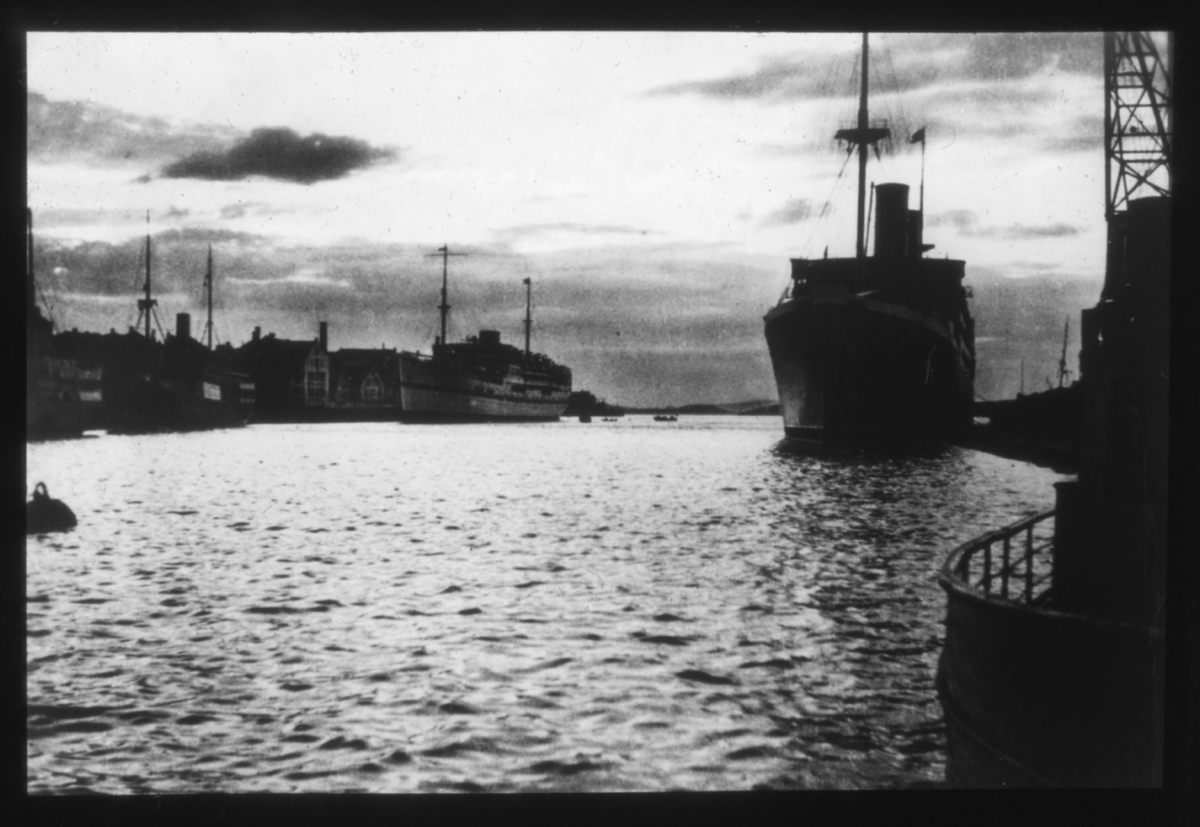 Dampskip ved kai i havn. To skip ligger til kai, skipet til høyre ser ut til å være et dampskip. Flere sjøhus ligger langs havneløpet til venstre.? En lossekran ses helt i kanten av høyre side av bildet. Begge skip har stor skorstein og ser ut til å være passasjerskip. Flere andre båter med mast ligger langs kai (skuter). Helt til venstre i bildet flyter det som ser ut til å være en fortøyningsblåse. Bildet er tatt i motlys og har høy kontrast.
 Muligens er motivet fra Trondheim eller en annen nordlig by?