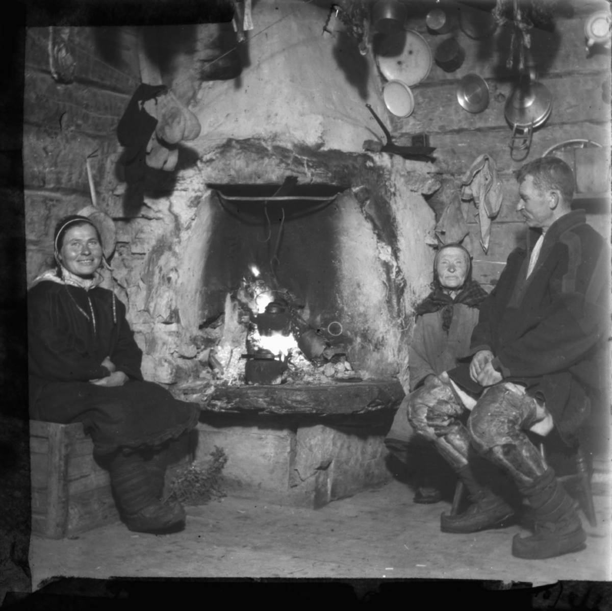 "Idyll ved gruen. G.15. 41" står det på glassplaten. Samisk famile sitter ved peis. Kaffekjellen henger over ilden. På veggene kan man se endel kjøkkenutstyr og på en snor henger det votter eller sokker til tørk. Alle har på seg samiske kofter, skaller på beina. Kvinnene har samisk lue. Mannen har også benklær sydd i skinn. Veggene er i tømmer. Peisen eller ildstedet i bakgrunnen.