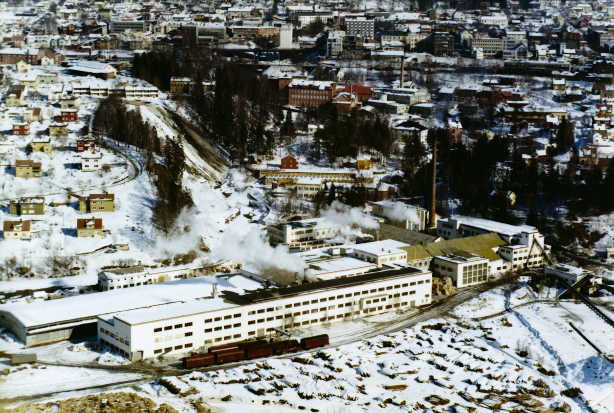 Lillehammer, Nedre bydel. Mesna Kartongfabrikk i forgrunnen med jernbanespor med flere vogner. Lagret tømmer. Stor, hvit bygningsmasse med fabrikkpipe. I bakgrunnen til venstre en bratt skråning som blir kalt "snøtippen". En stor, rødbrun bygningsmasse lenger bak med to fabrikkpiper er Gudbrandsdal Uldvarefabrikk.