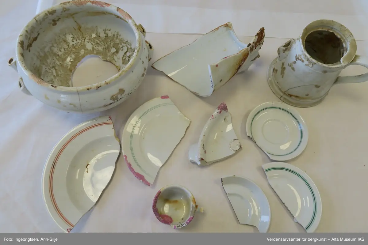 Bruddstykkene er av A) Terrin, B og C) Kanne, D) Dyptallerken, E) Flattallerken, F) Serveringsfat?, G og J) Tefat, H) Kaffekopp, I) Asjett. De er alle sammen hvite og noen av de har grønne eller røde striper rundt kantene. På undersiden av gjenstandene er det stempel med produsentnavn og hakekors med ørn over.