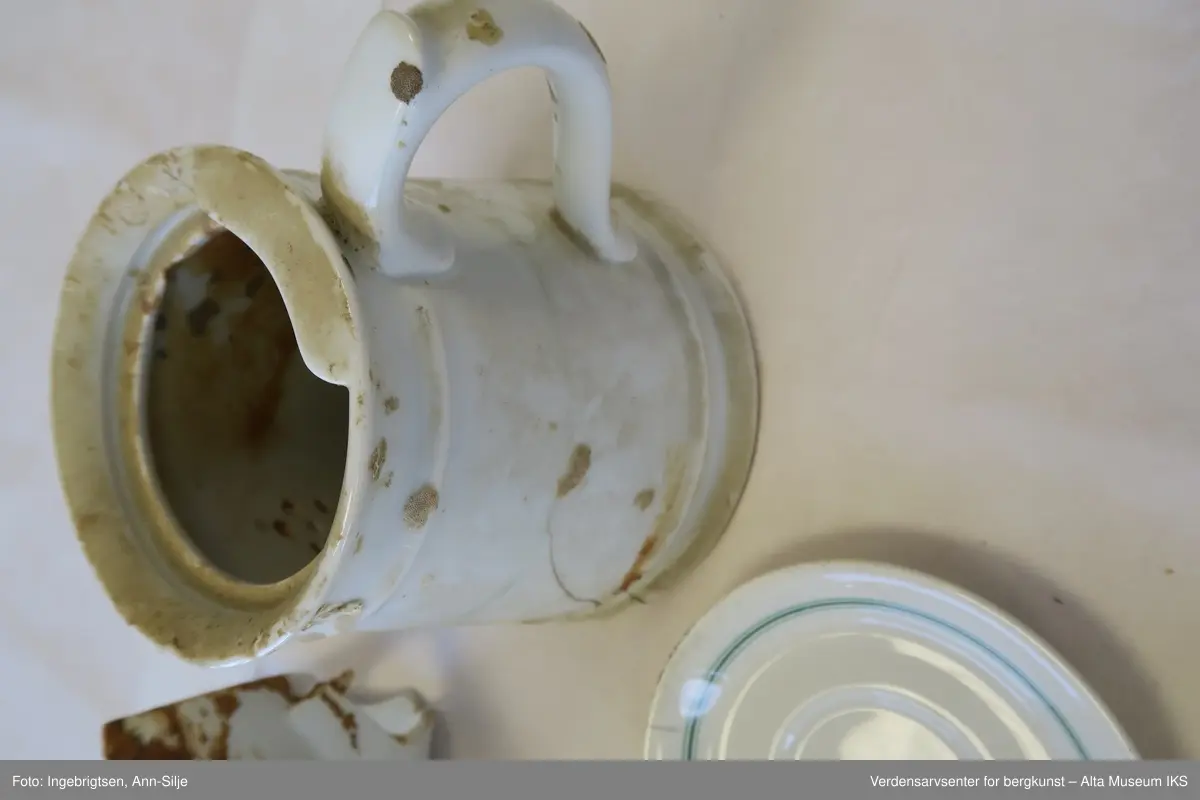 Bruddstykkene er av A) Terrin, B og C) Kanne, D) Dyptallerken, E) Flattallerken, F) Serveringsfat?, G og J) Tefat, H) Kaffekopp, I) Asjett. De er alle sammen hvite og noen av de har grønne eller røde striper rundt kantene. På undersiden av gjenstandene er det stempel med produsentnavn og hakekors med ørn over.