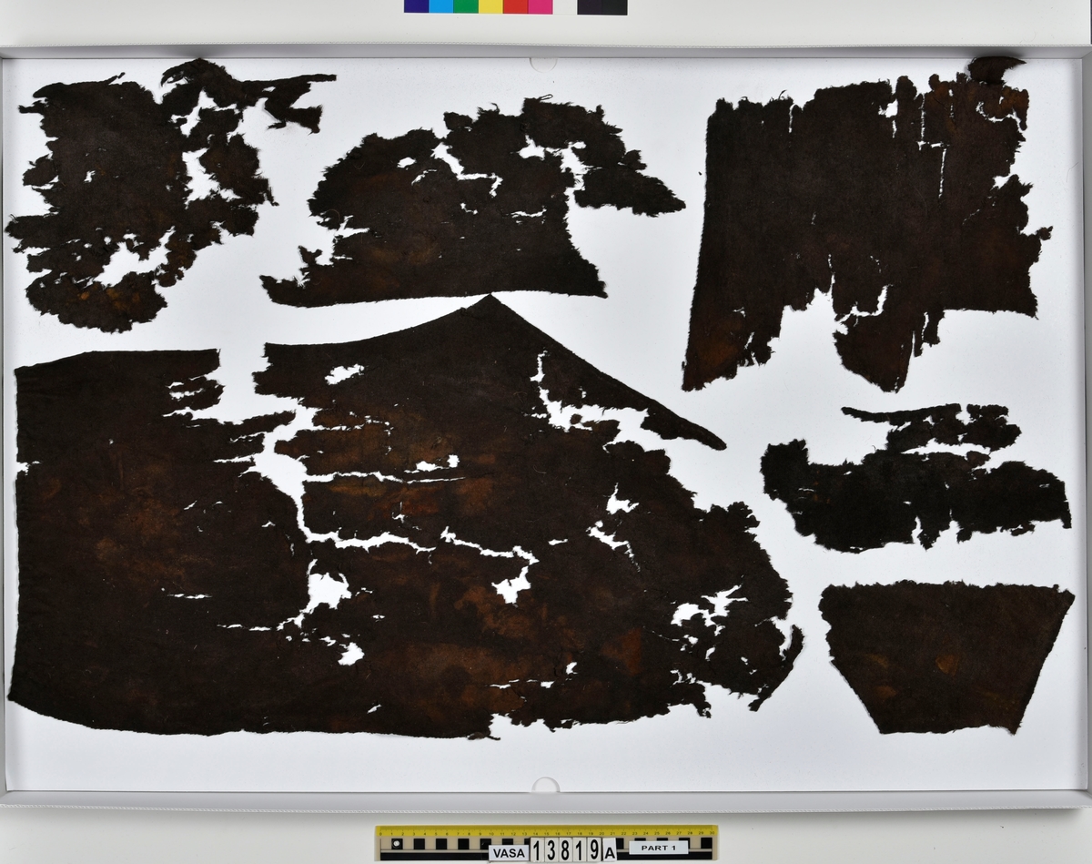 Textilfragment, byxa.
125 fragment uppdelade på fyndnummer 13819a-d.
Fnr 13819a består av 44 fragment av ull vävt i tuskaft och valkat på ena sidan. En del av fragmenten har originalkanter med sömmar och fållar.
Fnr 13819b består av 35 fragment av ull vävt i tuskaft. En del av fragmenten har originalkanter med sömmar.
Fnr 13819c består av 40 fragment av ull vävt i tuskaft. En del av fragmenten har originalkanter med sömmar. Fragmenten är mycket sköra.
Fnr 13819d består av 6 fragment mönstervävt band av ull.