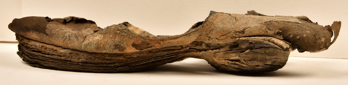 Sko.
Inuti skorna hittades även fragment från olika textilier som ursprungligen varit ett par strumpor. Idag sitter ett stickat fragment av ull fast i vänsterskon (23070) med avigsidan uppåt. I fyndet finns även en mängd lösa stickade fragment som idag är mycket korroderade och vissa helt mineraliserade av järn vilket har gjort textilen stenhård och därmed oerhört skör (23070a-c). Utifrån minskningar som några små fragment har så har det varit möjligt att rekonstruera strumpans tå. Genom att pussla ihop fragmenten som ursprungligen har suttit ihop har det på så vis gått att avgöra hur strumpans tå har varit konstruerad. Då textilen är så fragmentarisk är det inte möjligt att avgöra hur resten av strumpan har sett ut eller varit konstruerad. Däremot så finns det fragment av ett kypertvävt tyg av ull som på vissa ställen ligger dubbelt med det stickade 23070c). Ett av dessa fragment har stelnat i formen så som den suttit på fotens främre del inuti skon. Detta fragment täcker delvis det stickade tyget och har dessutom spår av kaststygn längs en kant. Så som det vävda tyget är placerat på det stickade tyget pekar på att man använt det som lagningslappar till sina stickade strumpor. 
I fyndet finns det även korroderade järnklumpar, ett tuskaftsvävt tyg av ull och rester av ett grovt bastfibergarn och två mindre läderfragment (23070d-f). Det är möjligt att även dessa har tillhört strumpan och/eller skon.

För mer detaljerad information från textildokumentationen (år 2022) se länkade filer.

Skodokumentation 2022:
Ovanläder. Tillverkad i getskinn av god kvalitet. Hör ihop med fyndnummer 23069 (Yngves skor). Tillverkades på en rak läst och bars på vänster fot. Inga dekorationer Det finns stygnhål som visar att det funnits invändiga förstärkningar (men förstärkningslappen saknas). Det finns delaminering men inga lagningar.
För mer information, se Länkade filer: Uppritning

Text in english:
This is the upper of a shoe of more “extreme fashion”, made of goatskin leather of good quality, probably made on a straight last and worn of the left foot, with no decorative features. The upper couples with 23069, together composing Yngve’s shoes. There are internal reinforcements missing from the upper and delamination, there are no repairs.
For more information, go to Länkade filer: Uppritning