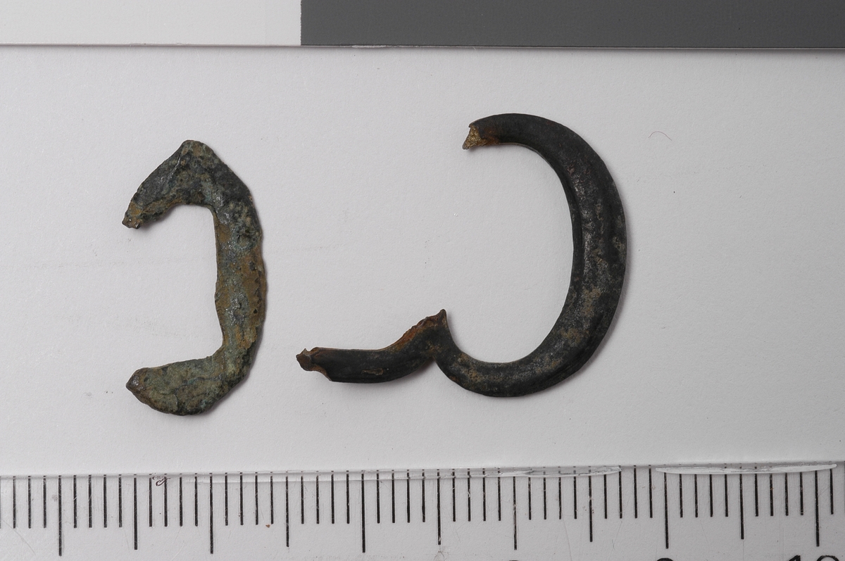 To små spenner av ulik størrelse og utforming.  Den minste er oval, med tynne, flate kanter og en rektangulær åpning i midten. Den større spennen har også oval utforming, oval indre rom og ser ut til å være en dobbeltspenne. Denne gjenstanden har også ovalt tverrsnitt på rammen. Ingen av de to spennene har bevart noe torn.