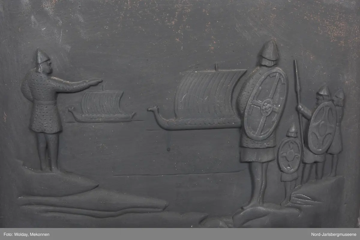 Vikingscene med fem vikinger og to skip, i front. PÅ toppstykket tre øksehoder på rad, vendt inn mot en  midtplassert medaljong med drageornament. Kokerommets kortsider relieff med vikingskip sett rett forfra for fulle seil.