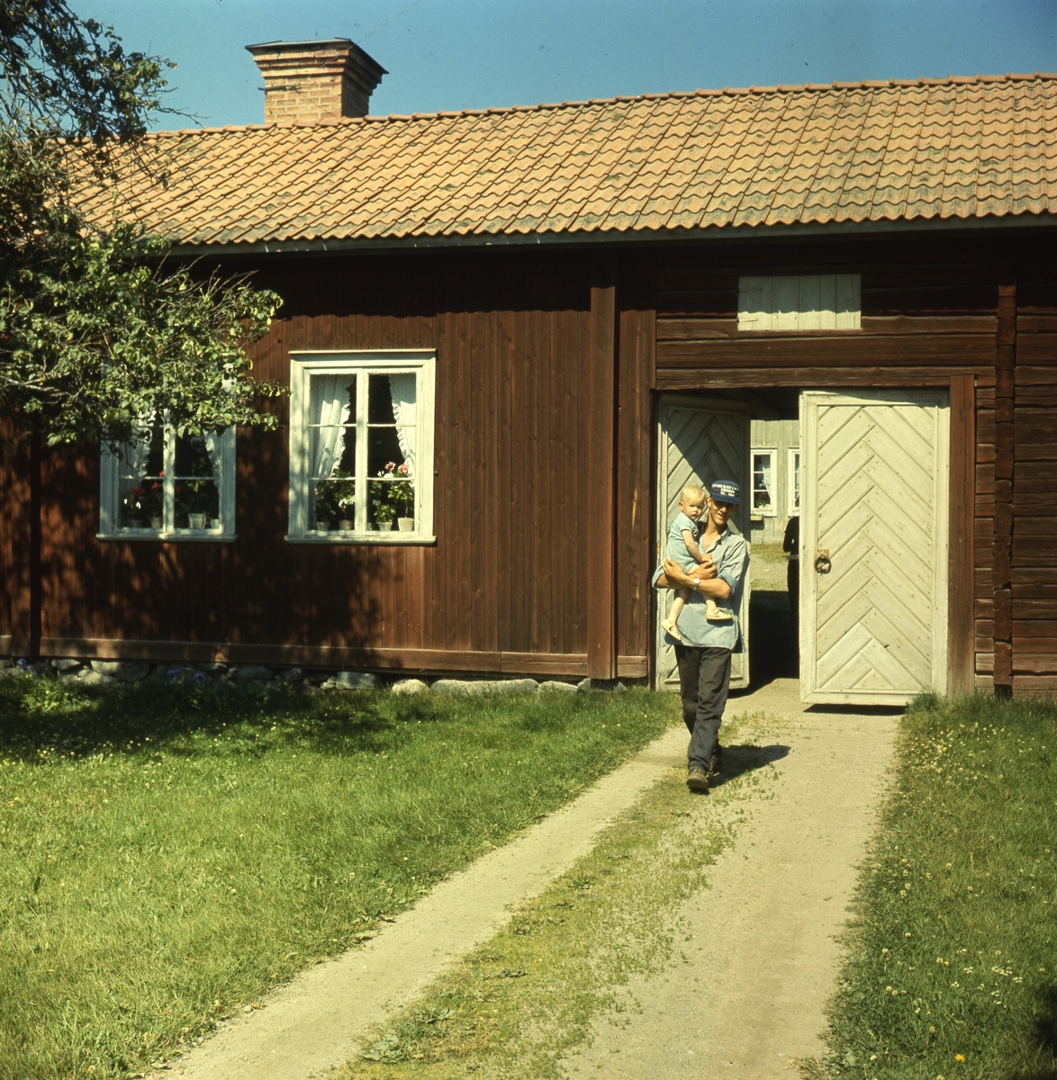 Portlidret vid Ol-Svens i Fors, Norrala. En man kommer ut från portlidret med ett barn på armen. Han går på vägens grässträng. Det är sommar.