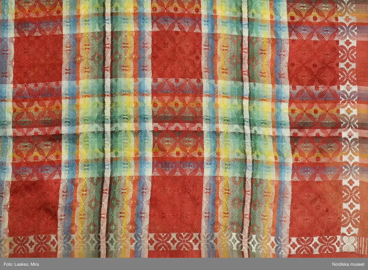 Kvadratiskt halskläde av siden, orangeröd botten med rutning i gult, grönt, ljusblått och vitt samt en smal ytterbård med stiliserad blomma längs 2 sidor i vitt och längs de andra i gult. Botten mönstervävd i ett bindemönstermönster med kryss och små stjärnor, i ytterkant utanför bården slät tuskaft. Uppsrispad kort frans runt alla sidor. I ena hörnet en rund svart hallstämpel med texten. Stockholm 1825 och i mitten St. Eriksbild. Troligen vävd i en skaftvävstol.
Typen var vanlig på 1820-talet och finns från flera håll i landet bl.a 98530 från Äppelbo och 93669 från Malung  i Dalarna. även 44279 från Västmanland och 91632, 152104, 162623 från Småland.
I fint skick
/Berit Eldvik 2011-11-23