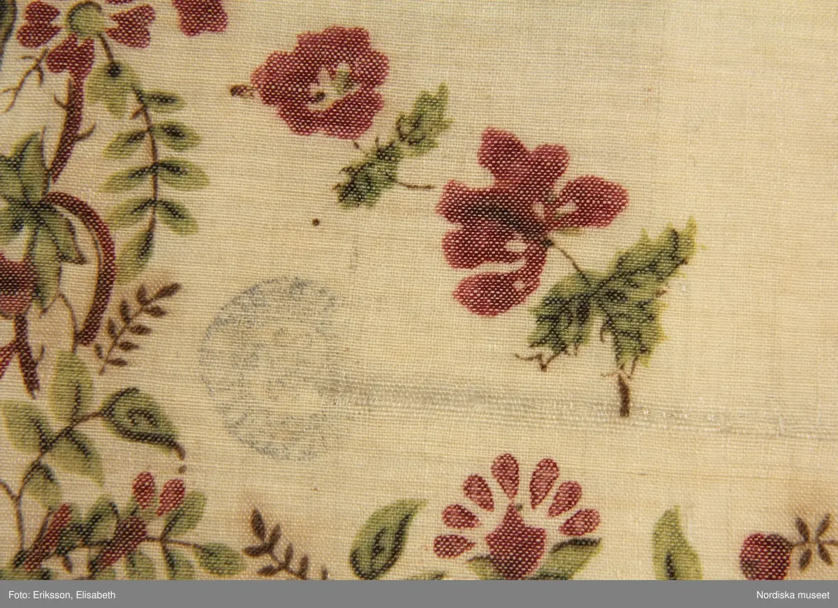 Kvadratiskt halskläde  av gråvit tuskaftad yllemuslin med tryckt mönster i matt lila , grönt och något brunt.  I mittspegels strödda  små blomkvistar.  I bården smal blomslinga  i lila och grönt med tunna bruna stjälkar.  Runtom gles iknuten frans av vitt ullgarn. I ena hörnet en rund svart stämpel med texten: Göteborgs hallstämpel.
Anm. fransar saknas, små fläckar och hål.
De tunna fina muslinschalarna  med tryckt mönster kom in i det folkliga modet som brudschalar vid mitten av 1800-talet.  En del var importerade men tillverkning fanns också i Göteborg.
 Tryckaren J.J.Amelung i Stockholm var den första som kunde trycka på tunt ylletyg 1834. Han gjorde dukar med mönster efter de sk. kashmirschalarna. Se Henschen: Kattuntryck , 1992 sid 70-71.
/Berit Eldvik 2012-01-05