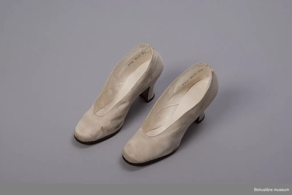 Högklackad damsko, utvändigt vitt siden, invändigt vitt skinn, lädersula. Pumps storlek 38.
Skrapmärken och missfärgningar. 
Greta Karlssons brudskor från år 1922.
Placerade i skolåda UM018555.

Se UM018527
Sammanhör med UM018591 Brudslöja, UM018595 Brudbukett och UM018596 Diadem.