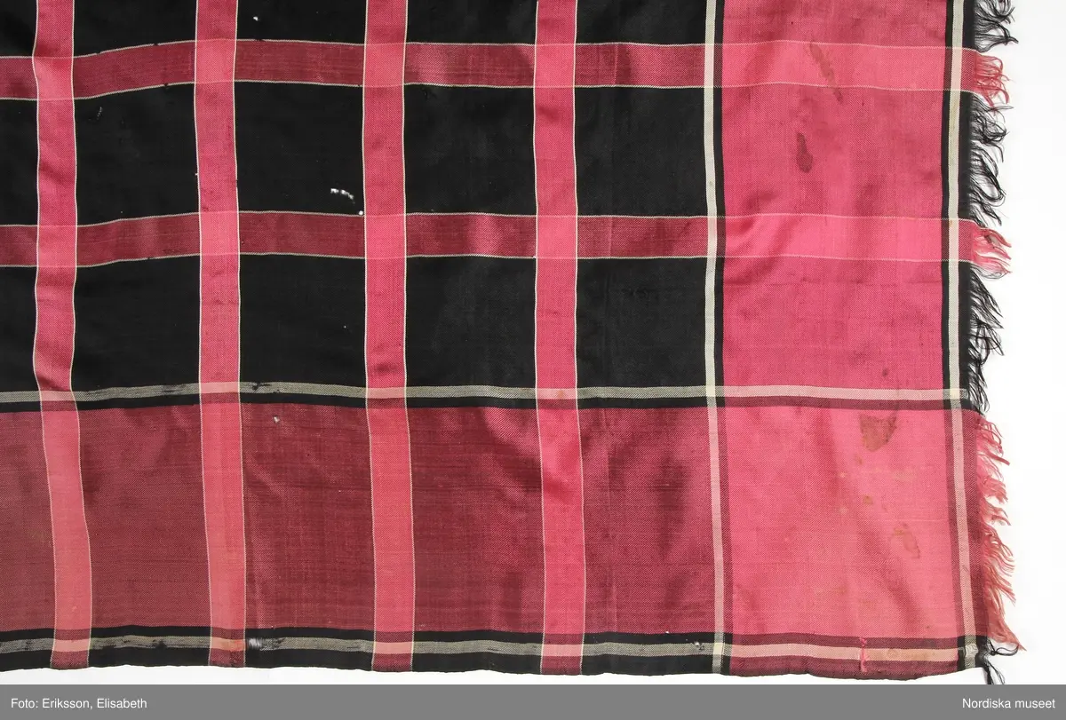 Kvadratiskt halskläde av siden i kypert. Svart botten med inrutning med rosa ränder med vit kontur och 13 cm bred kantbård i samma rosa färg,  runtom alla sidor upprispad kort frans.
Anm. Något blekt  i 2 av hörnen samt några fläckar
/Berit Eldvik 2011-11-16