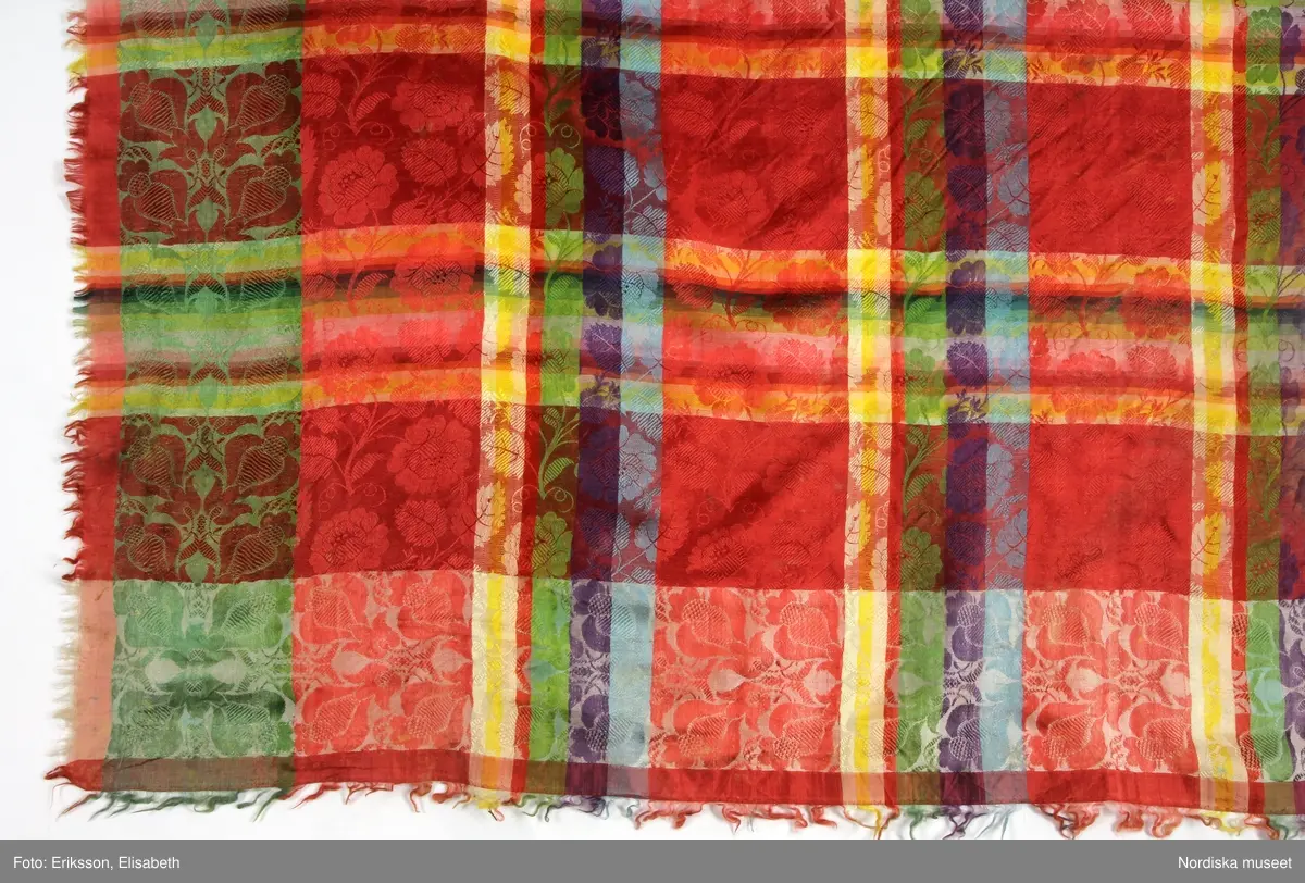 Kvadratiskt halskläde av siden, klarröd botten med rutor i grönt, ljusblått, ljuslila och gult med ett yttäckande invävt mönster, troligen med jacquard, med slingrande rosor. Kantbård med "päronlika" blommor/frukter som på 2 mötande sidor är gröna och på de 2 andra vita. Upprispad kort frans på alla sidor.
I ena hörnet en tydlig svart rund stämpel med texten: Stockh. hallstemp 1839.
Anm. Stor fläck i två av hörnen.
/Berit Eldvik 2011-11-11