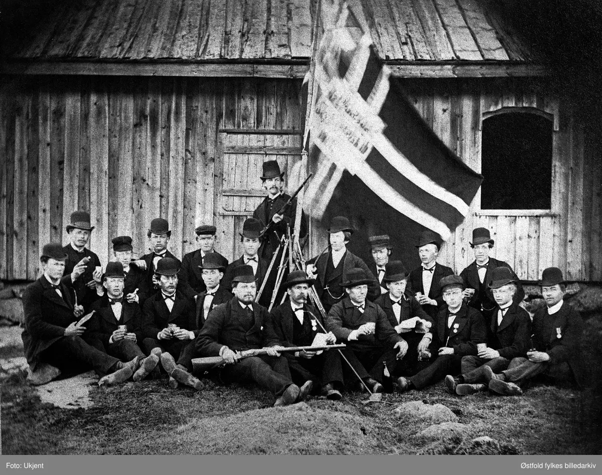 Rakkestad skytterlag og medlemmer av Eidsberg skytterlag  foran skytterhuset på Søndre Fladstad i Rakkestad i 1880 (1880-åra). I lagets historie er det oppgitt at bildet er fra skytterhuset på Schiesmyren. Det er Rakkestad Skytterlags flagg (med sildesalat) som vaier i bakgrunnen. Skytterlaget ble stifta 17. mai i 1861 med navnet Rakkestads Skydeselskab. Skytterhus ble oppført i 1863, og bane anlagt. 
På bildet er det også skyttere fra Eidsberg skytterlag som gjester skytterlag i Rakkestad.. Karene koser seg med en dram og er pynta med medaljer, det kan være et stevnearrangement.
Eidsberg skytterlag blei stifta 17. mai 1858 på Mustorp. 

Navneopplysning fra nesten identisk motiv publisert på Oslo Museums side: "Bak, nr. 3 fra høyre er verkseier Hans Martin Torp. Foran nr. 3 fra høyre er K. E. Jahren. (Kristian Edvard Jahren)". Begge fra Rakkestad.

Bygdebok for Eidsberg 1940. s. 451, har oppgitt disse navnene for samme bilde,  altså ant navn på de som var fra Eidsberg (?): 
"Mannen lengst til venstre er Ole Torkildsen Filtvedt. De to i midten foran - med geværet over knærne - er en folkehøyskolelærer (enten Peder Velde (f. 1860) eller Lasse Trædal f. 1857 - må være P. Velde) og Ingebrigt (?) Præsthus (med medalje på brystet). Lengst til høyre sitter Johannes Spestad. Bak til venstre sitter Nils Granberg, og nummer to fra høyre i bakerste rekke er Anders Torp fra Trømborg."