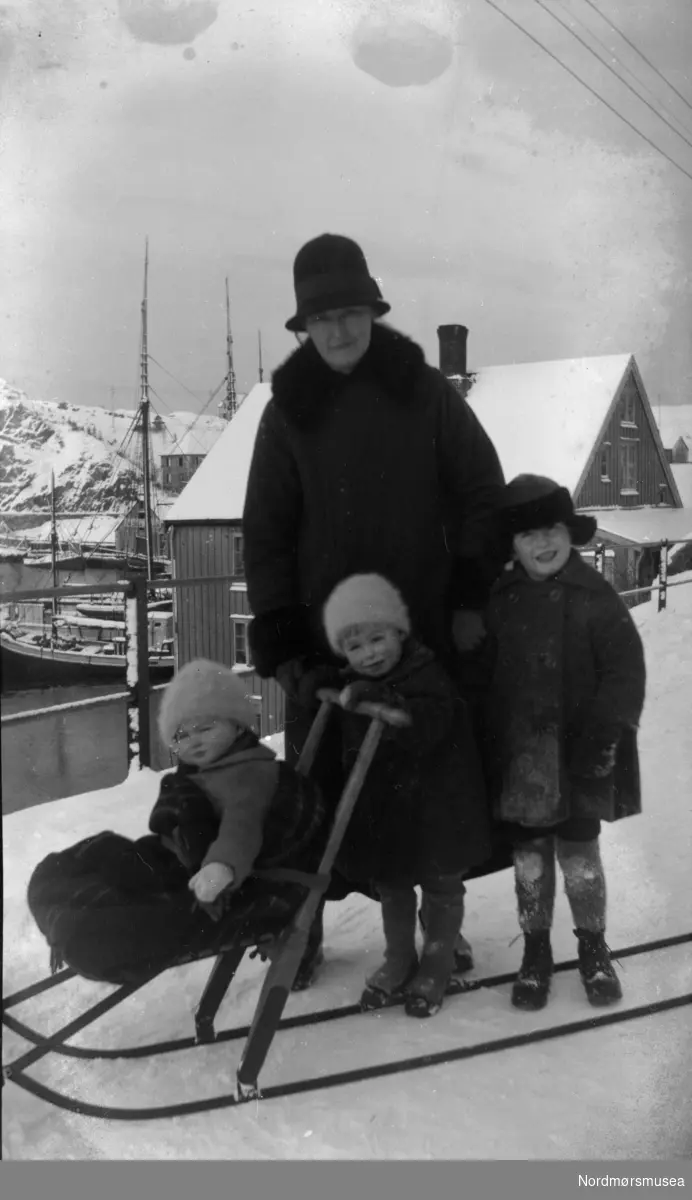 Foto av en kvinne ute med tre små barn med spark. Datering er usikker men trolig rundt 1917-1920. Bildet ser ut til å være fotografert langs Kranaveien i Vågen på Kirkelandet i Kristiansund. Fra Nordmøre museums fotosamlinger.