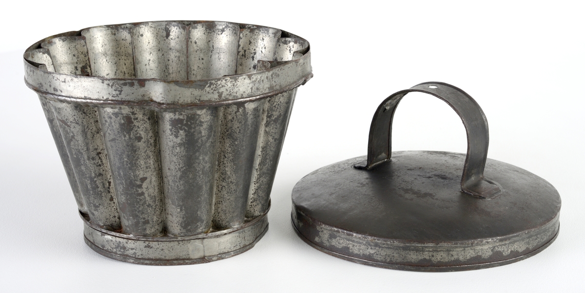 Form med lokk brukt til enten kakebaking eller brødbaking. Formen er rund og har 12 riller på innsiden og 12 tunger rundt kanten. Oppe på formen er det et bånd som holder formen sammen (kun dekor). I bunnen av formen er det en sirkulær fordypning samt en fordypning som forestiller en blomsterstilk og et blomsterblad. Ved hvelving av kaken/brødet vil dette fremstå som et opphøyet mønster på toppen av kaken. Lokket er svart utenpå og grått inni. Lokket har en hank der det er et hull (til oppheng?).