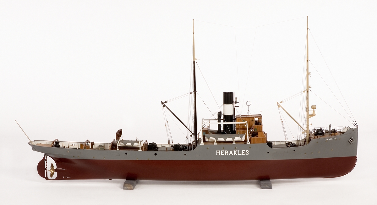 Fartygsmodell av bärgningsångaren HERAKLES. Hel i block av trä. Gråmålat skrov med rak stäv och häckakter, långt akterdäck med fyra bogserbågar. Flera pumpar och vinschar på däck. Svart skorsten med vitt bälte.