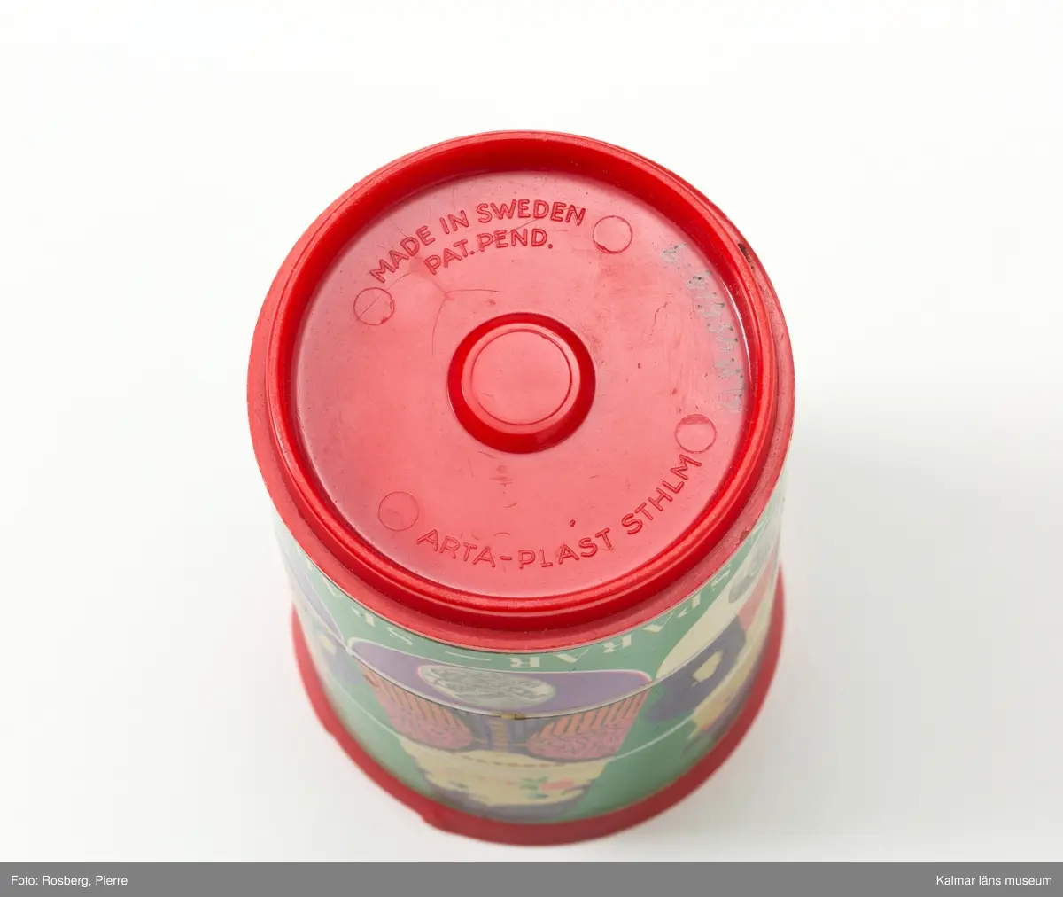 KLM 45519:9 Sparbössa. Rund sparbössa i röd plast. På ovansidan präglat i plasten, Sparbanken, den gamla sparbanksloggan med en ek samt, familjebanken. Här finns också en springa att stoppa pengar i. Sidorna består av tre olika rader som kan vridas i olika lägen. De olika raderna har en klistrad pappersremsa med motiv. Den översta raden har ansikten av människor från olika länder t ex Sverige, England och Kina. Mittdelen utgör människornas torso och nedre delen visar mynt från respektive land. På den nedre delen står, Hela världens sparar – spara för din värld. I botten, präglat i plasten,  MADE IN SWEDEN PAT. PEND. ARTA-PLAST STHLM”.