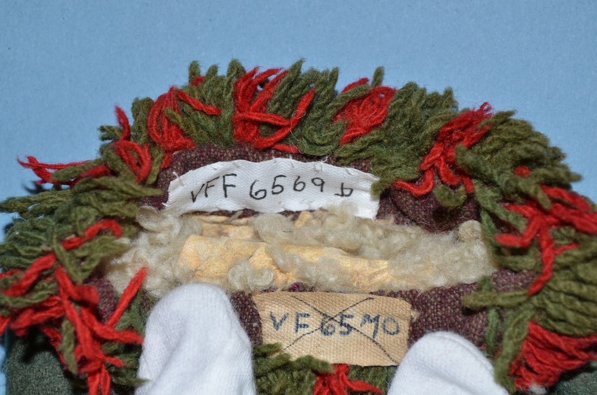Vott (a+b), kypertvevd ull, grønn. Forside tommel mørkere grønn og stampet. Håndsydd. Rød og grønn frynsebord i kantene, festet på kant av toskaft bomull (brun og beige). Fôr i lammeskinn.