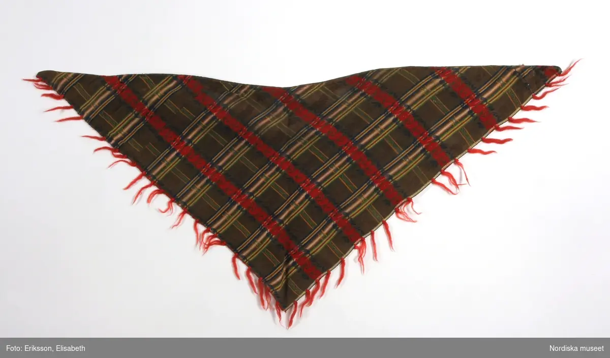 Trekantigt halskläde av troligen jacquardmönstrad bomullssatin.  Grönbrun botten randig i  ljusare grönt, gult och blått med invävd bladslinga i bottenfärgen. Tvärgående mönsterbårder i rött och blått  samt smalare i gult och grönt, vilket ger ett rutigt  tyg. Tvärbårderna i inslagsflotteringar.   Tyget har 2 materialskarvar. I ytterkanterna iknuten gles frans av rött ullgarn.Fodrad till större delen med ett grön/brunrutigt bomullstyg, snibben bak ofodrad.
 Tillverkad av delar från ett kvadratiskt halskläde.
/Berit Eldvik 2012-02-09