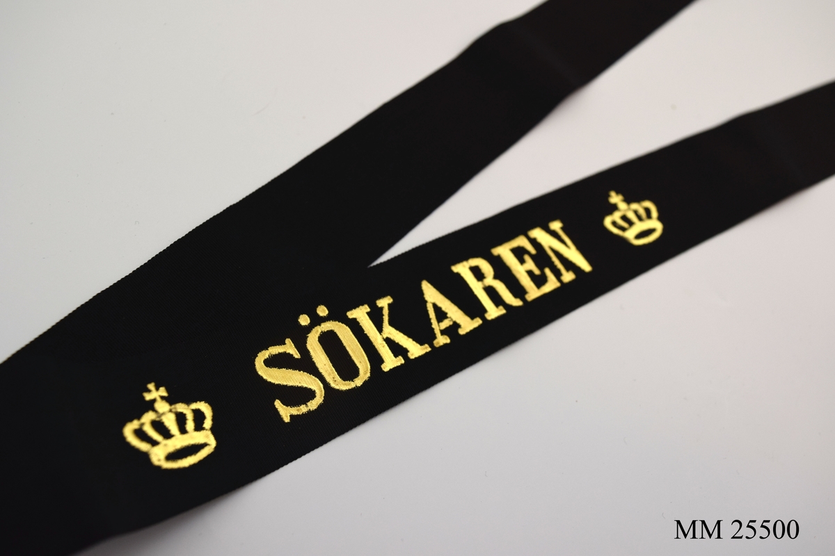 Mössband av svart sidenrips. Guldfärgad text, "Sökaren", med två kronor på vardera sida.