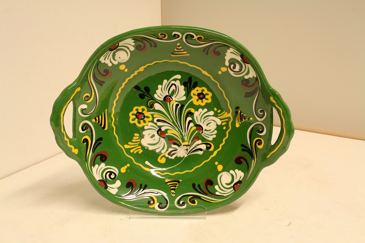Avlangt, avrunda fat med håndtak i hver ende. Fatet er grønt med rosemaling-lignende blomstermotiv i hvitt, gult rødt og svart.