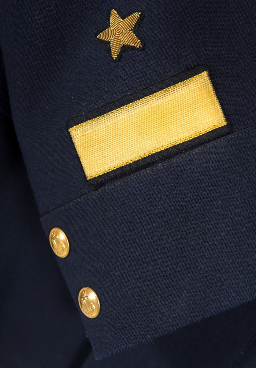 Kolett från ryska marinen för kapten av första rangen. Blått kläde. Fodrad med svart siden, i ärmarna vitt siden. Knäppt med 5 gulmetallknappar. 2 bröstfickor med lock och 2 höftfickor med lock. 1 ficka på var sida i fodret. 2 gulmetallknappar på var ärm. 1 guldstickat sterck och 1 guldstickad stjärna på var ärm. Axelklaffar med 3 längsgående guldgaloner mellan dessa svart silke, knapp i gulmetall 3 stjärnor i vitmetall.