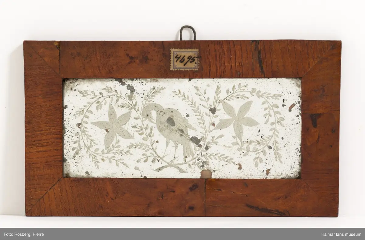 KLM 4695. Spegel, prydnadsspegel. Ram av trä med faner. Spegel med glasetsning med motiv i form av en stor fågel (örn?) med böjd hals som flankeras av stjärnlika blommor med sirliga rankor med små blad på. Enkel, platt, polerad rand.