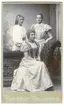 Porträtt på tre kvinnor i klänningar. Sittande är Thyra Berg, fotografbiträde hos Therése och Lisa Wallgren mellan 1897-1901, samt även systerdotter till dessa.