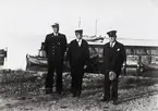 Tre herrar, varav en uniformsklädd, stående vid hamnen på Revsudden.