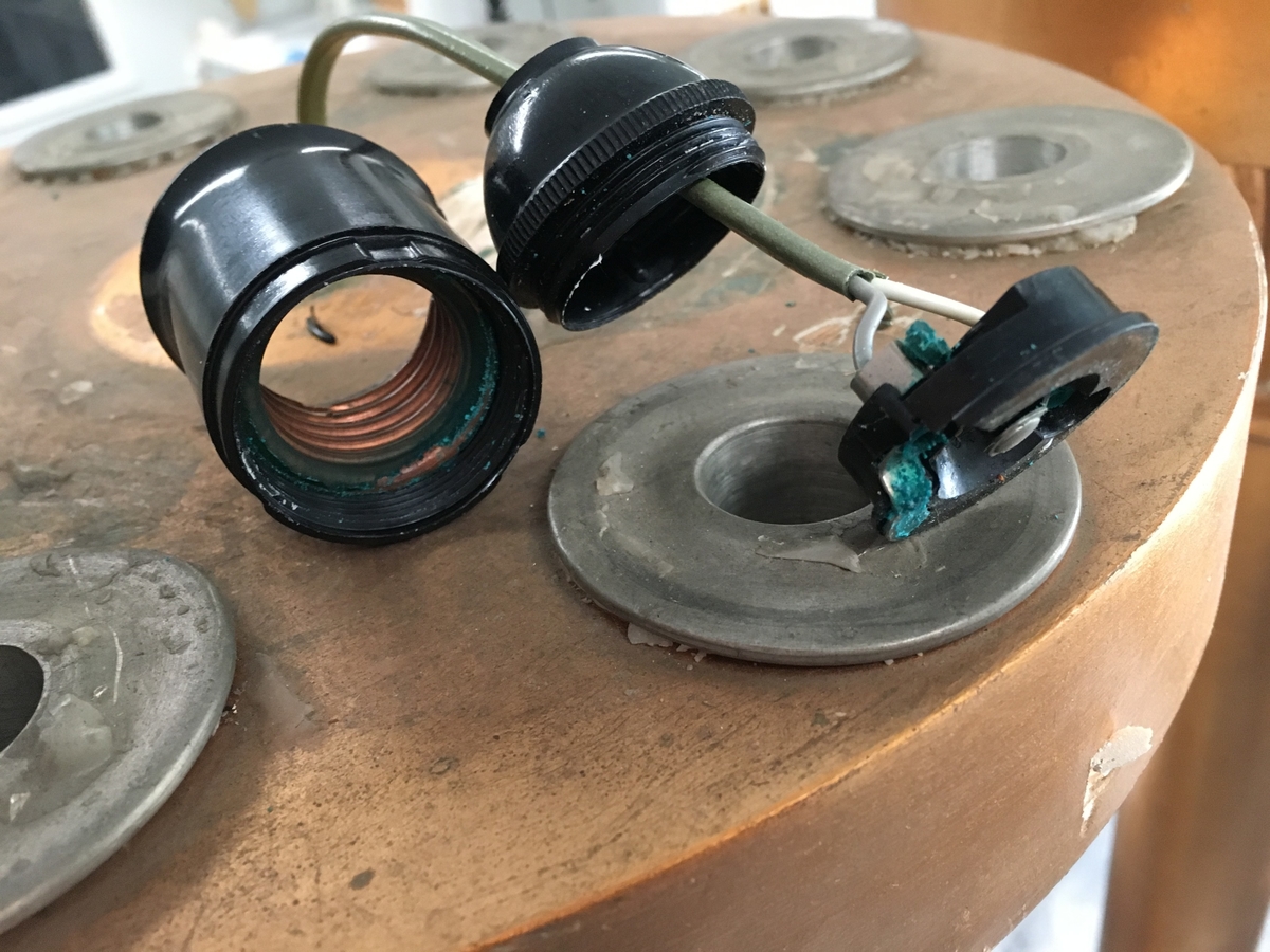 Forgylte gulvstående lamper for flere lys, både stearinlys og elektrisk. Hull til stearinlys langs kanten på hele topp-platen. En åpen lampeskjerm i glass omkranser det elektriske lyset midt på abakus, med bølgende kant (som en fakkel). Lampen er formet som en frittstående gresk søyle med base. Glatt utforming, med et område med bladutskjæringer like under midten, samt på kapitélet. Rund abakus og firkantet base. 
To av lampene har et forgylt tresylinder rundt pipen for det elektriske lyset. 
Var sannsynligvis ikke el.belysning originalt.