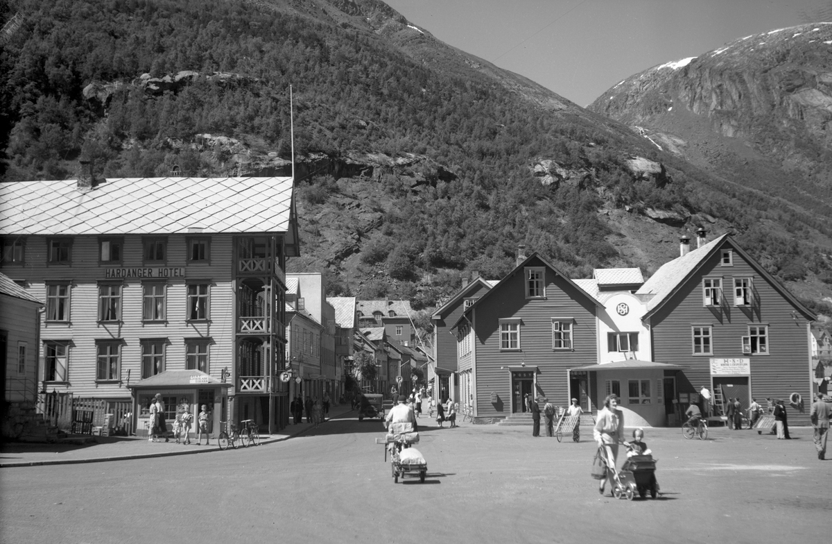 Odda torg, Hardanger Hotel, Torget 2-4, Brotateigen, Dagestad sin kiosk med plakaten: Frukt, sjokolade, tobakk, hotell, gate, postkontor, HSD-ekspedisjon, HSD-emblem, butikkar, mann på sykkel med vogn, kvinne med barn og barnevogn, mann med handkjerre, syklar, folkeliv, bil, sommardag i Odda.