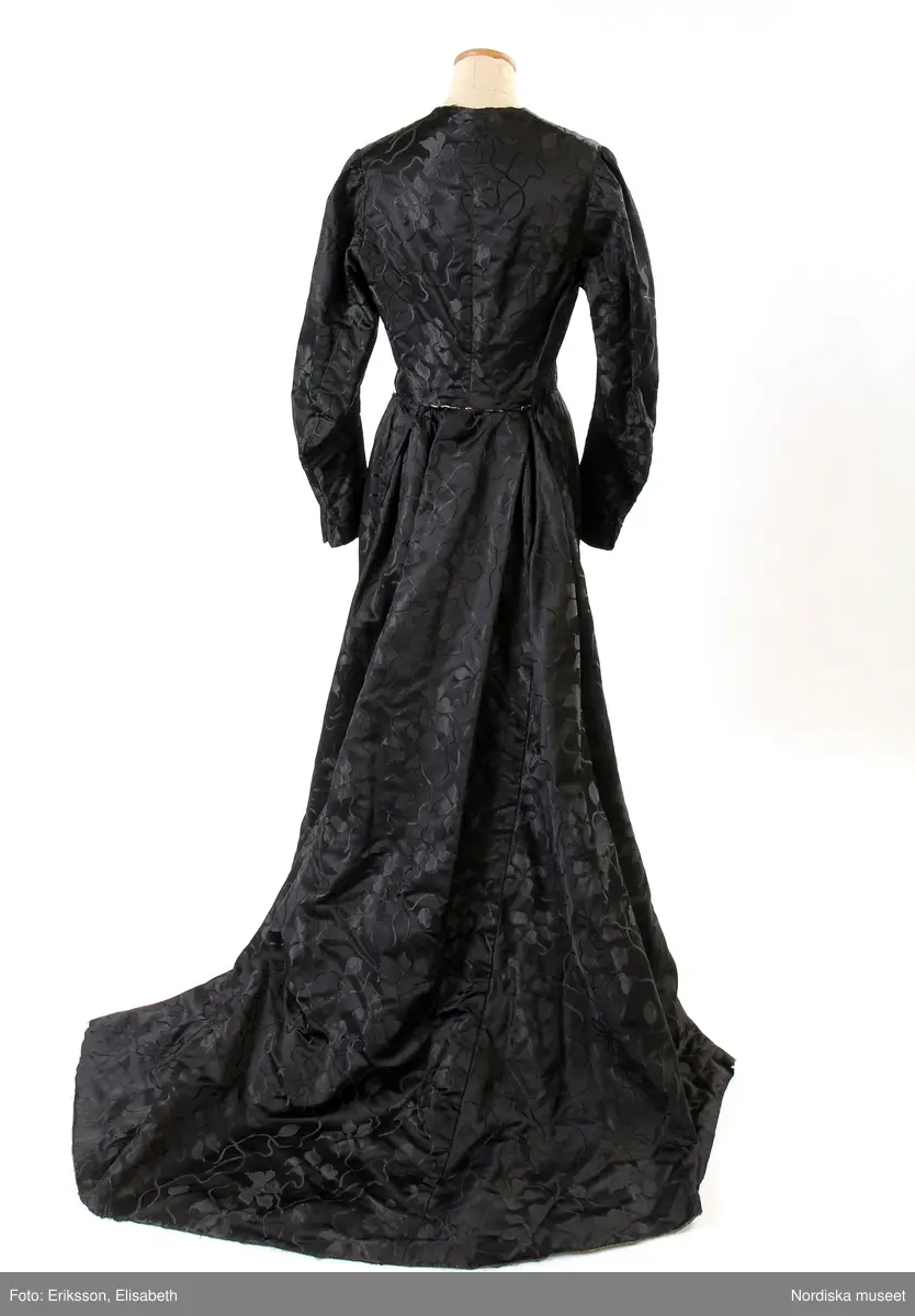 Högtidsklänning med släp, svart jacquardvävt siden, med mönster av blomstergirlanger. Långärmat klänningsliv med fyrkantig urringning. Antydan till fårbogsärm med lätt rynkning vid axelpartierna. Söm mitt på ärmen som kan tyda på att den förlängts från att från början ha varit en 3/4-delsärm. Klockformad kjol, med flera våder, två sömmar på framdelen. Linning ca 2 cm. Liv och kjol är fästade med hakar, men även delvis ihopsydda. Kjollängd fram 105 cm, bak 130 cm. Ärm 60 cm. Midjemått ca 66 cm, ryggmått, livstycke 41 cm. Slitage på utsatta ställen, som nedre kanten och vid linningen. Delvis omsydd. Tryckknappar för knäppning av kjoldelen.
/Karin Dern 2017-05-18