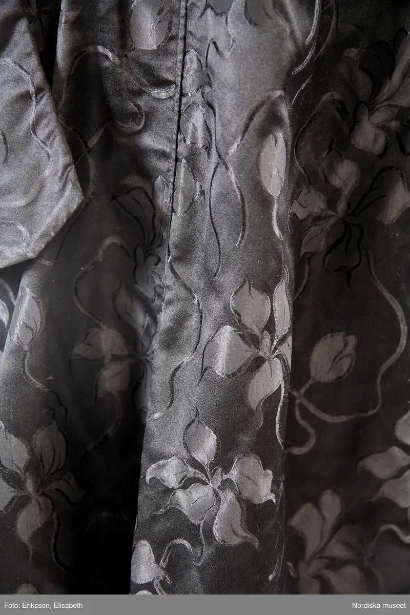 Högtidsklänning med släp, svart jacquardvävt siden, med mönster av blomstergirlanger. Långärmat klänningsliv med fyrkantig urringning. Antydan till fårbogsärm med lätt rynkning vid axelpartierna. Söm mitt på ärmen som kan tyda på att den förlängts från att från början ha varit en 3/4-delsärm. Klockformad kjol, med flera våder, två sömmar på framdelen. Linning ca 2 cm. Liv och kjol är fästade med hakar, men även delvis ihopsydda. Kjollängd fram 105 cm, bak 130 cm. Ärm 60 cm. Midjemått ca 66 cm, ryggmått, livstycke 41 cm. Slitage på utsatta ställen, som nedre kanten och vid linningen. Delvis omsydd. Tryckknappar för knäppning av kjoldelen.
/Karin Dern 2017-05-18