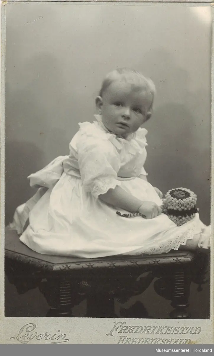 atelierfotografi av liten gut i kvit kjole som sit på bord og held ei rangle i handen