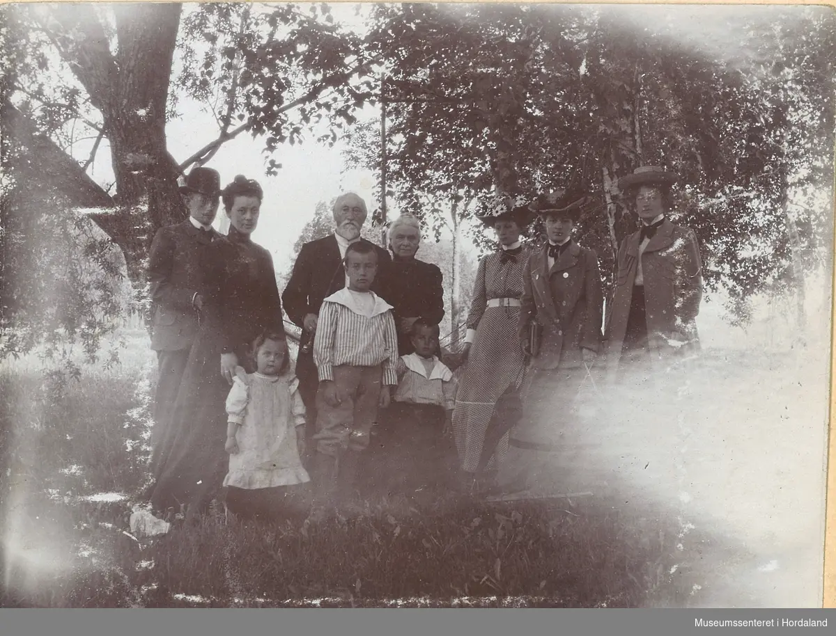 amatørfotografi av eit eldre par med tre unge kvinner til høgre for seg, ungt par til venstre og tre born. Dei står under store trær på ein trapp i hagen