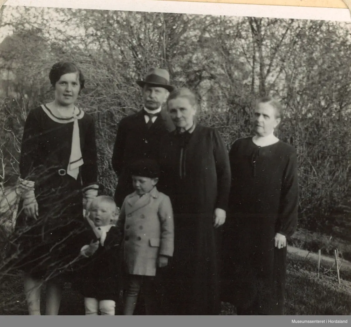 amatørfotografi av eldre ektepar med svigerdotter, to barnebarn og ei anna kvinne  ute i ei hage med busker og trær rundt seg