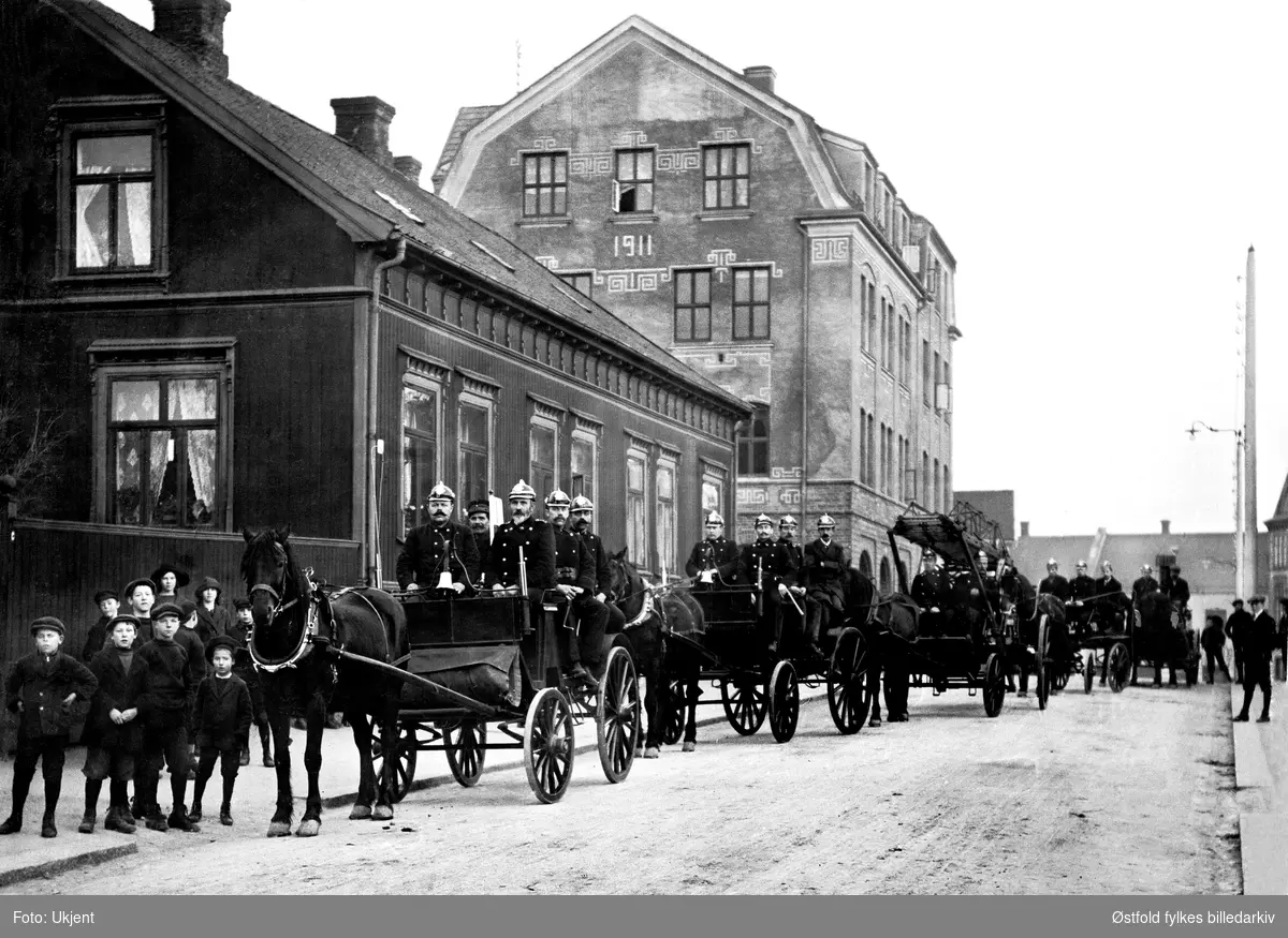 Brannkorpset i Sarpsborg  oppstilt med hester og vogner og uniformerte brannkonstabler i Sigvat Skaldsgate utenfor brann- stasjonen. Barneflokk til venstre. Stort bolighus til venstre. Murbygning 1911 - brannvakta i bakgrunnen. Bildet tatt etter 1911.