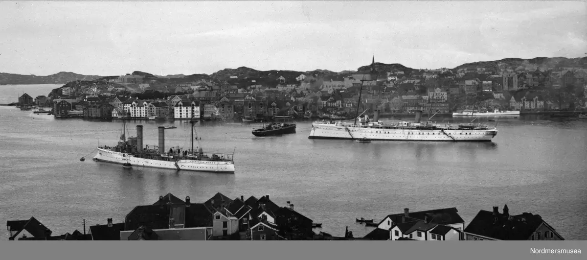 "Christiansund N." "Chr. sunds havn" Foto av flere store fartøy i Kristiansund. Trolig fra et flåtebesøk. Ranheimsæter antas å være fotograf basert på fotografens stempel på KMb-2017-021.0601. Bildet er stemplet med årstallet 1903, så bildet er fotografert dette året eller de påfølgende hvis bildet er montert på en "utdatert" papplate.  - Det store fartøyet omtrent midt i bildet er det tyske keiserskipet S.M.S. Hohenzollern, bygget i 1893. Opprinnelig hadde skipet karakteristiske skorsteinskapper som ble fjernet under moderniseringen av maskineriet i 1906.  Til venstre i bildet ligger en lett krysser av Gazelle-klassen oppankret. Det hvitmalte fartøyet langs kai til høyre i bakrunnen er trolig SMS Sleipner, torpedobåten S97 ombygget til følgeskip for Hohenzollern. De øvrige bildene i denne serien viser at S.M.S. Hohenzollern har de karakteristiske skorsteinskappene (som på dette bildet er knapt synlige), og at bildene derfor er tatt før 1906. (Info fra Espen Arnestad 2017)  Fra Nordmøre museums fotosamlinger.