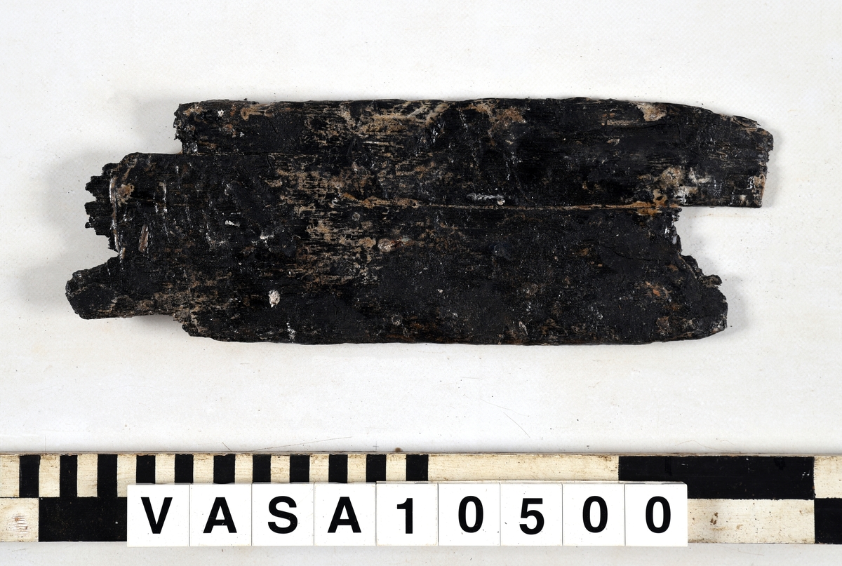 Bräda med fästanordning vid ena kortsidan, möjligen del av möbel. Vittrad, nästan ingen originalyta kvar. Tjockare i ena änden. Avbruten i båda ändar, stor spricka längsmed föremålet samt många repor och mindre sprickor. Saknas uppgift om salthalt.