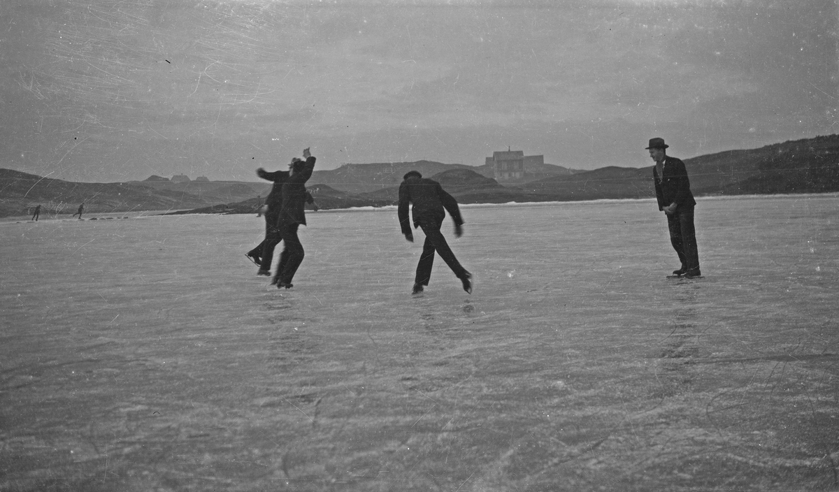 Vinter. Fire menn har det moro på skøyter på isen. Hus og land i bakgrunnen. Hverdagsfoto.