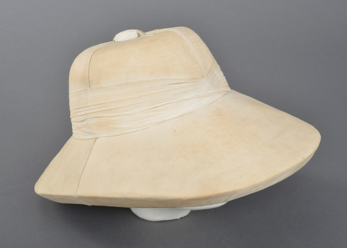 Hatt trekt med stoff. Pullen er sydd sammen av fire deler. Trekt knopp på toppen. Rundt pullen er hatten dekorert med flere lag med stoff. Pullen og bremmen er av lett og tykt materiale. Pullen er foret og i toppen er det trykt fabrikkmerke. Bremmen er foret med grønt stoff. Skinnkant er festet i nedere kant av pullen.