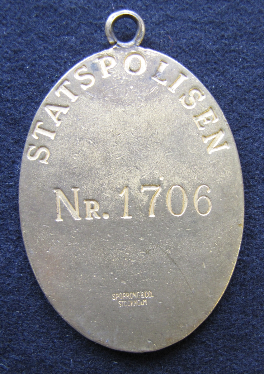 Polisbricka, oval med en ögla i toppen. 

På framsidan: Lilla riksvapnet, dvs en krönt sköld med tre kronor. Bakom skölden eklöv och korslagda fasciner. Under emblemet står det POLIS

På frånsidan: STATSPOLISEN står den i det övre kanten. Mitt på står NR. 1706. Även tillverkaren Sporrong & CO, Stockholm finns ingraverat.