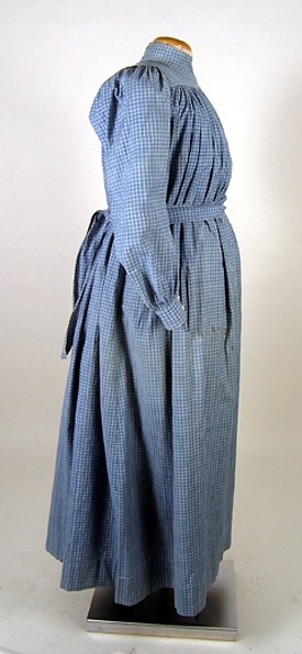 Klänning (bakförkläde) av blå-vit rutigt bomullstyg. Klänningen är rynkad runt om det släta oket. Oket är fodrat med ett vitrandigt bomullstyg. Manschetterna har sprund och knäpps med 2 trädda knappar. Framtill på höger sida finns ett hål för en ficka. I midjan finns en dragsko som knyts framtill på magen på insidan. Klänningen knäpps baktill med 7 hakar och sydda hyskor. Ett 11 cm brett skärp i samma tyg knytes runt midjan. Fläckar framtill samt ett hål.