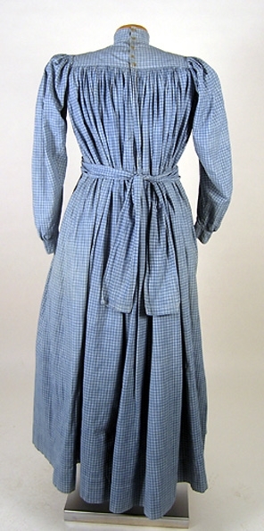 Klänning (bakförkläde) av blå-vit rutigt bomullstyg. Klänningen är rynkad runt om det släta oket. Oket är fodrat med ett vitrandigt bomullstyg. Manschetterna har sprund och knäpps med 2 trädda knappar. Framtill på höger sida finns ett hål för en ficka. I midjan finns en dragsko som knyts framtill på magen på insidan. Klänningen knäpps baktill med 7 hakar och sydda hyskor. Ett 11 cm brett skärp i samma tyg knytes runt midjan. Fläckar framtill samt ett hål.