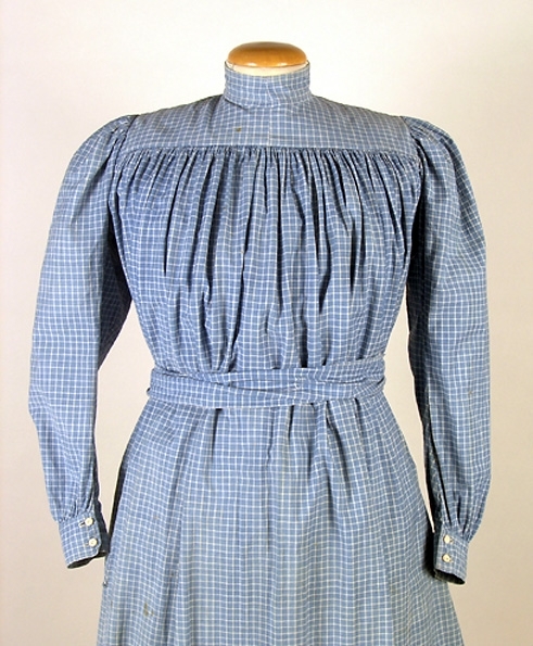 Klänning (bakförkläde) av blå-vit rutigt bomullstyg. Klänningen är rynkad runt om det släta oket. Oket är fodrat med ett vitrandigt bomullstyg. Manschetterna har sprund och knäpps med 2 trädda knappar. Framtill på höger sida finns ett hål för en ficka. I midjan finns en dragsko som knyts framtill på magen på insidan. Klänningen knäpps baktill med 7 hakar och sydda hyskor. Ett 11 cm brett skärp i samma tyg knytes runt midjan. Fläckar framtill samt ett hål.